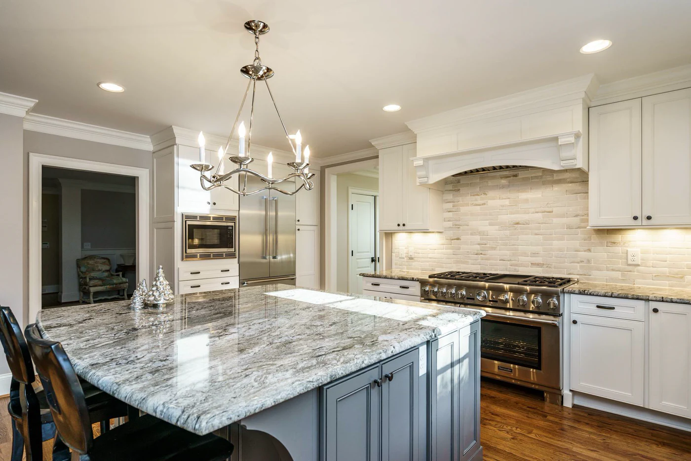 Kitchen island with a gray-and-white stone top and gray painted base under a polished nickel chandelier, with a stainless pro range and white cabinets behind
