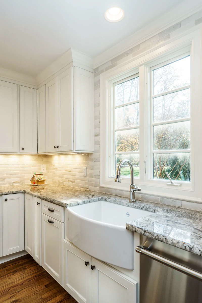 White farmhouse sink under a double window, with stone countertops, white cabinets and under-cabinet lighting on a light tile backsplash