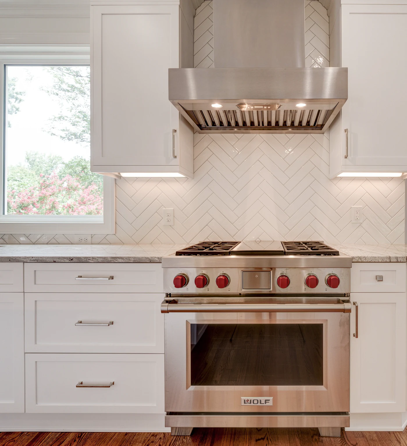 Stainless Wolf range with red knobs under a stainless hood, white herringbone tile backsplash, white shaker cabinets and a window beside it