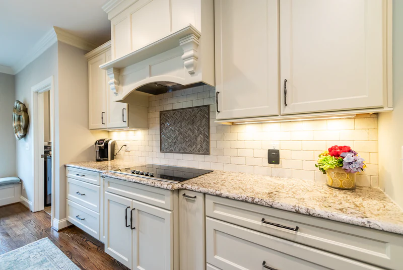 Cream cabinets with a wood range hood with corbels, a herringbone mosaic tile inset, a black glass cooktop and warm under-cabinet lighting on a stone counter