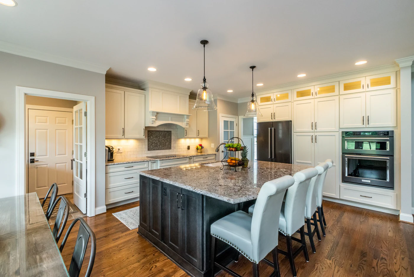 Dark-stained kitchen island with a speckled stone top and four light upholstered stools under glass bell pendants, cream cabinets and wall ovens behind