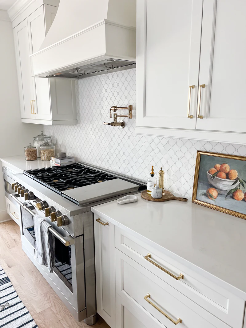 White range wall with a painted wood hood, arabesque tile, brass pot filler and brass cabinet pulls, stainless range with brass knobs
