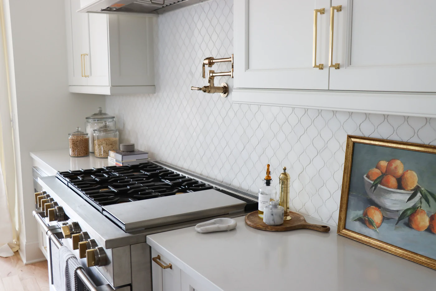White arabesque tile backsplash with a brass pot filler above a stainless range with brass knobs, white cabinets with brass pulls