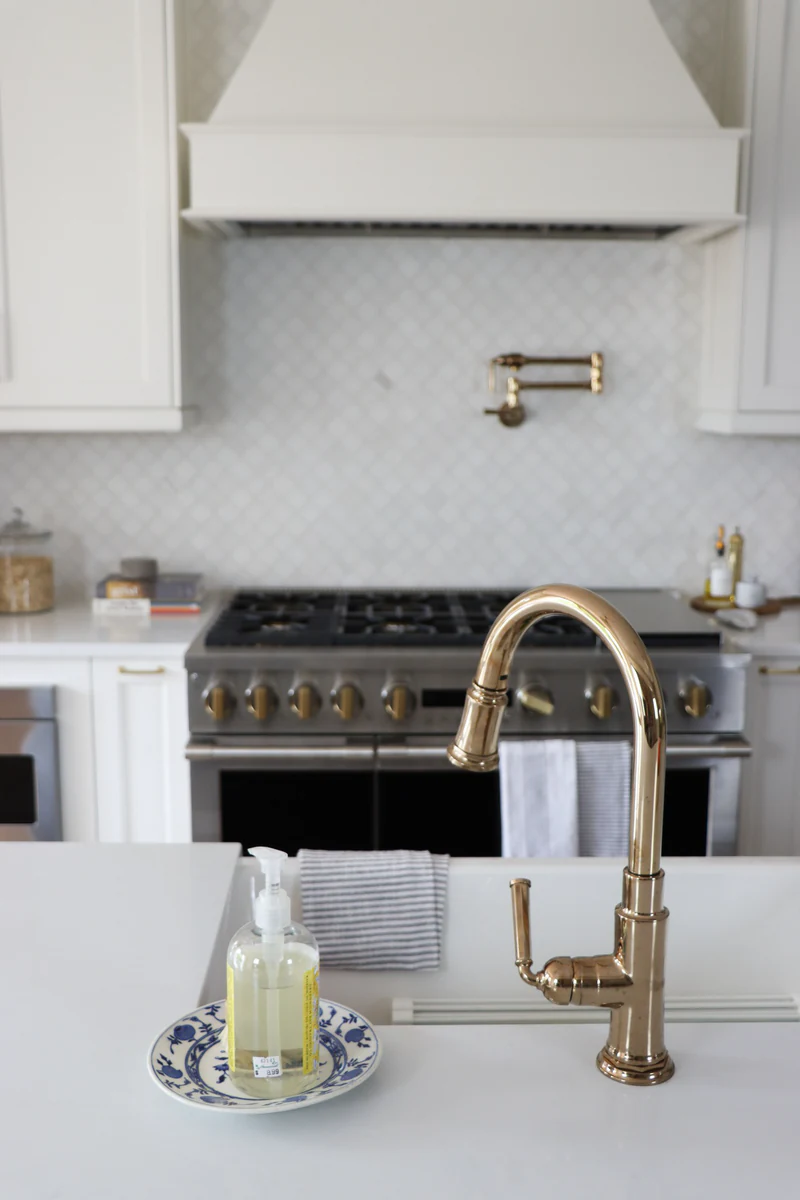 Polished brass faucet at an island sink, with a stainless range with brass knobs, a white hood and arabesque tile behind