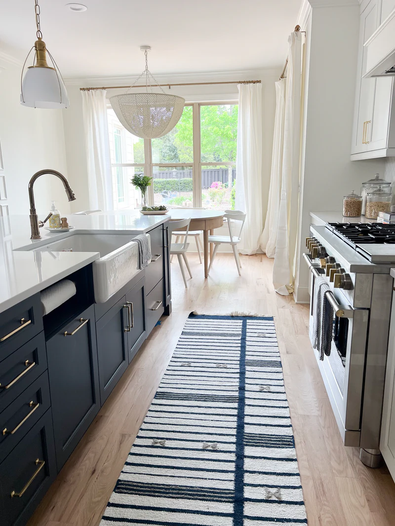 Navy island with a white farmhouse sink and brass pulls, a striped runner, and a stainless range facing a breakfast nook with a round table