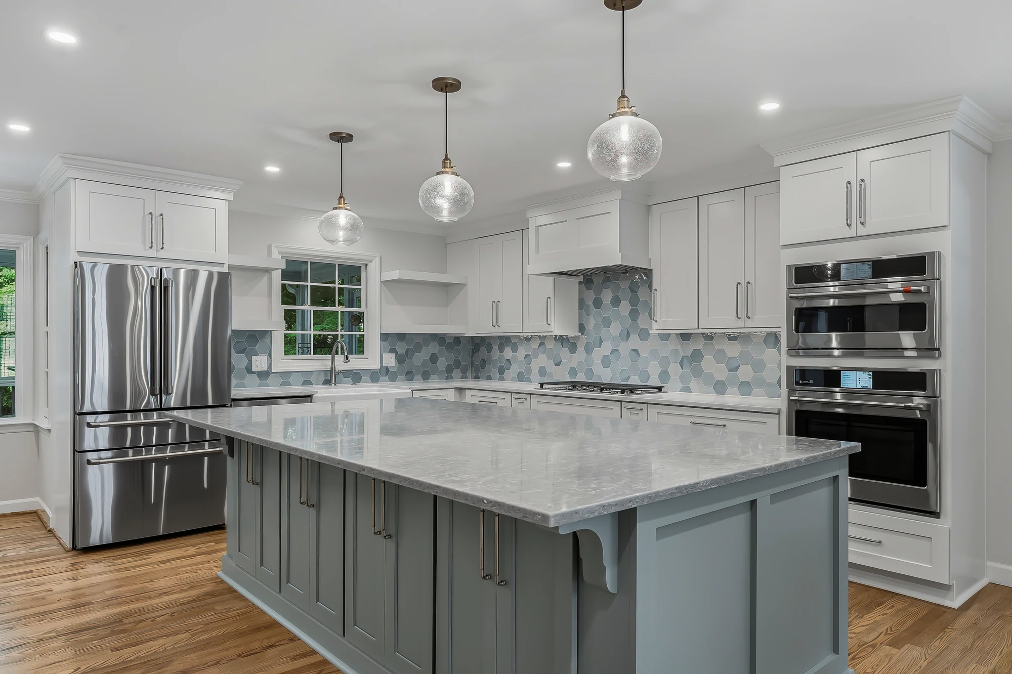 White kitchen with a large blue-gray island, gray stone top, three glass globe pendants, blue hexagon tile and stacked wall ovens
