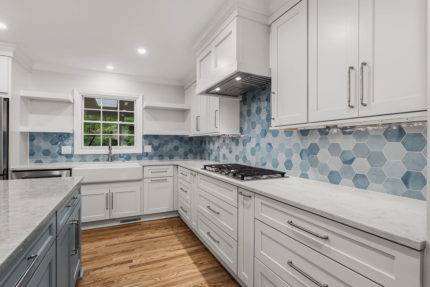 White shaker cabinets wrapping a U-shaped kitchen with a blue hexagon tile backsplash, gas cooktop, farmhouse sink under a window and open shelves