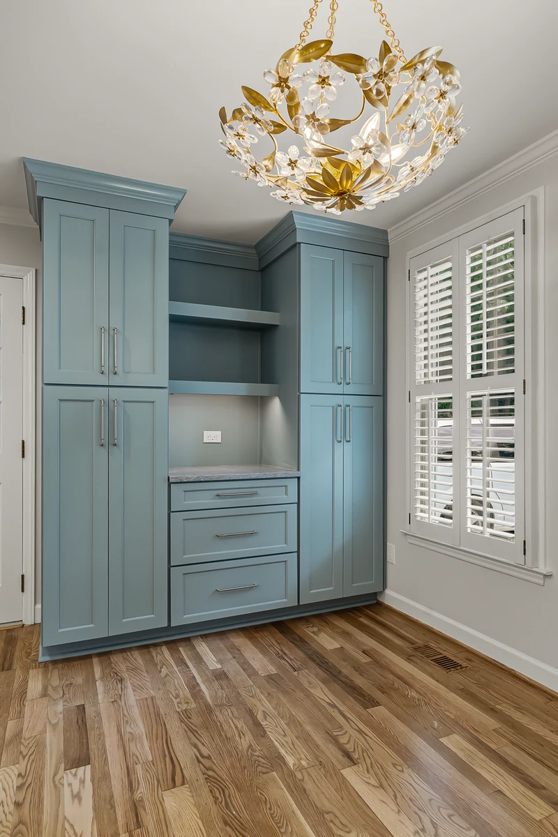 Blue-gray built-in cabinet wall with open shelves and drawers, a gold floral crystal chandelier above, and a window with white plantation shutters